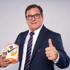 Uśmiechnięty mężczyzna w garniturze, z piłką w ręku i kciukiem w górze. Wygląda jak trener reprezentacji Polski, gotowy do akcji.