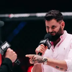 Wojtek Gola, znany z FAME MMA, udziela wywiadu. Uśmiechnięty, w różowej koszuli, z mikrofonem w dłoni.