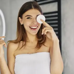 Uśmiechnięta kobieta w ręczniku wykonuje demakijaż oczu wacikiem.