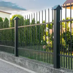 Czarna, metalowa furtka w prawie, z ostrymi zakończeniami, otacza zadbany ogród z kwitnącymi krzewami i cyprysami.