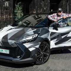Man lutar sig mot en Toyota C-HR Hybrid i kamouflagemönster. Bilen är parkerad på en kullerstensgata framför en byggnad med en automat.