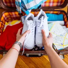 Comment ranger des chaussures dans une valise : une paire de baskets blanches est tenue au-dessus d'une valise ouverte remplie de vêtements et d'une carte.