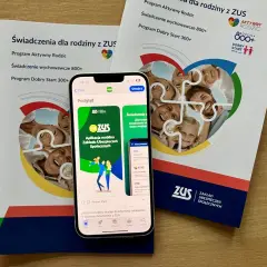 Aplikacja mZUS na telefonie pokazuje świadczenie wychowawcze 800+ dla rodziny. Ulotki ZUS informują o programach dla rodziców.