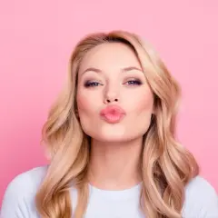 Donna con capelli biondi e trucco labbra naturale, che manda un bacio su sfondo rosa.