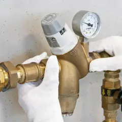 Mani guantate regolano un riduttore di pressione con manometro, mostrando come si regola la pressione dell'acqua.