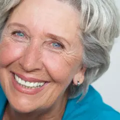 Donna anziana con capelli grigi e occhi azzurri, sorride mostrando le rughe intorno alla bocca, segno di una vita vissuta con gioia.