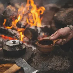En hand håller en träkopp med kaffe vid en lägereld. En kaffepanna står bredvid, redo att koka kaffe på gammalt vis.