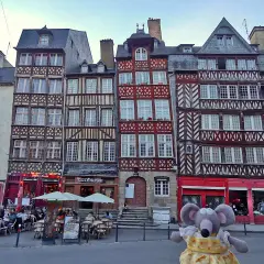 Une souris en peluche pose devant des maisons à colombages colorées, un aperçu de ce que faire à Rennes.