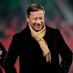 Uśmiechnięty Dariusz Banasik w czarnym płaszczu i beżowym szaliku, na tle trybun stadionu.