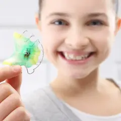 Niña sonriente muestra un retenedor dental verde y amarillo, listo para limpiar.