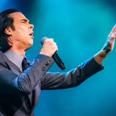 Nick Cave sjunger passionerat med mikrofonen i handen under en nick cave tour. Hans arm är utsträckt mot en blå bakgrund.