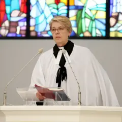 Kobieta w stroju duchownego, z białą narzutą i czarną kryzą, stoi przy pulpicie. To pastor, który przygotowuje się do wygłoszenia kazania.