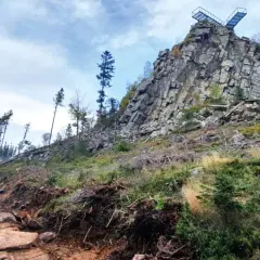 Kamienista ścieżka prowadzi ku skalistemu szczytowi z platformą widokową. W oddali widać las, a krajobraz przypomina ten z **Trójaka w Lądku Zdroju**.
