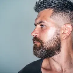 Mann mit Bart und tiefer fasson haarschnitt, im Profil auf grauem Hintergrund.