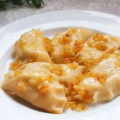 Pyszne pierogi ruskie, polane skwarkami z cebulki, na białym talerzu.