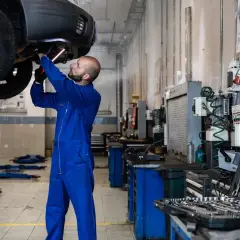 Mécanicien en bleu travaillant sous une voiture soulevée, outil à la main. Il pourrait s'agir d'une étape pour comment nettoyer un fap démonté.