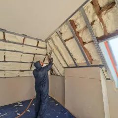 Un ouvrier en combinaison projette de la mousse polyuréthane pour isoler un grenier. L'isolant blanc recouvre les poutres et les murs.