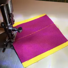 Close-up of a sewing machine needle and foot over purple fabric with a yellow border, demonstrating how to thread a sewing machine.