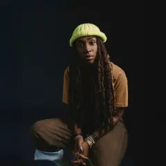 Jackie Hill Perry, mit Dreadlocks und einer neongrünen Mütze, sitzt nachdenklich vor dunklem Hintergrund.