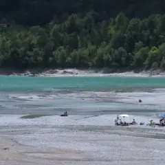 Persone rilassate sulla spiaggia del lago di Barcis, con acque turchesi e boschi sullo sfondo.