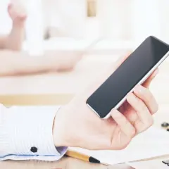 Dłoń trzyma smartfon, być może przeglądając ofertę kredyt orange. W tle widać biurko i inne osoby.
