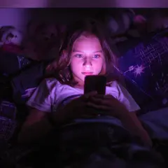 Dziewczynka leżąca w łóżku, patrząca w ekran telefonu w ciemności.