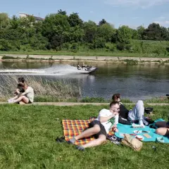 Majówka nad rzeką w Polsce: ludzie odpoczywają na trawie, a na wodzie ścigają się skutery wodne. Idealne miejsce, by spędzić wolny czas.