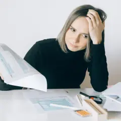 Kobieta zamyślona nad dokumentami, z ręką na czole, przy biurku ze stertą papierów.