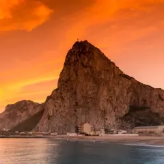 Gibraltar, czy to państwo, majestatycznie wznosi się nad morzem o zachodzie słońca. Pomarańczowe niebo maluje dramatyczne tło dla skalistej skały.