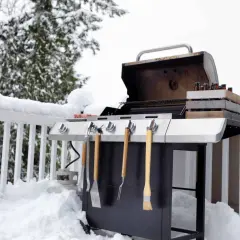 Un barbecue d'hiver prêt à l'emploi, avec des bouteilles de bière dans une caisse en bois et des ustensiles. La neige recouvre la terrasse et la balustrade.