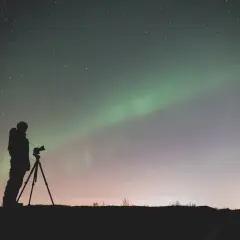 Sylwetka fotografa z aparatem na statywie pod rozgwieżdżonym niebem i zorzą polarną. Efekt bokeh gwiazd dodaje magii.
