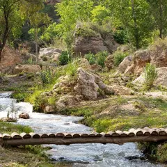 Un puente rústico cruza un arroyo en un paisaje rocoso y verde, evocando la serenidad de una cascada en Chelva.