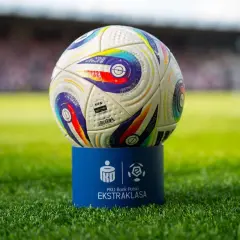 Piłka do piłki nożnej na niebieskim postumencie z logo Ekstraklasy na tle stadionu.