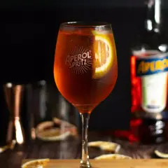 Szklanka Aperol Spritz z plasterkiem pomarańczy, butelka Aperol i inne składniki na drewnianym stole.