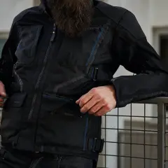 Mężczyzna w czarnej kurtce motocyklowej Rebelhorn Scandal II z niebieskimi akcentami, oparty o metalową barierkę.