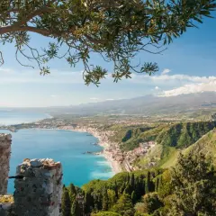 Vista panoramica di Naxos, Sicilia, con il mare azzurro, la costa e l'Etna innevato sullo sfondo, incorniciata da un ramo d'ulivo.