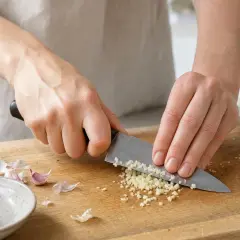 Mani che tritano finemente l'aglio su un tagliere di legno, esaltando le sue proprietà benefiche in cucina.