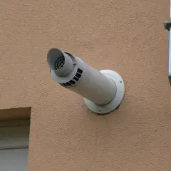 Boczny wyrzut spalin przez ścianę budynku z jasną elewacją, obok metalowego kanału wentylacyjnego.
