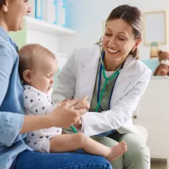 Uśmiechnięta lekarka bada niemowlę, które siedzi na kolanach mamy. Wykonuje test na odwodnienie, delikatnie dotykając rączki dziecka.