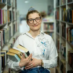 Bibliotekarka z okularami trzyma stos książek. W tle regały pełne literatury. Czy wakat co to znaczy?