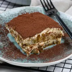 Ein Stück cremiges Tiramisu ohne Alkohol, bestäubt mit Kakaopulver, auf einem blauen Teller.