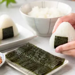 Dłonie formują pyszne onigiri. Obok miseczka z ryżem, nori i posypką.