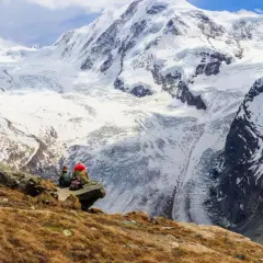 Two hikers rest on a rocky outcrop, gazing at the majestic, snow-covered peaks of the tour de Monte Rosa.