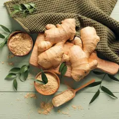 Imbir na co pomaga? Świeże korzenie i mielony imbir na drewnianym tle, otoczone zielonymi liśćmi.