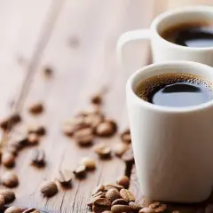 Dwa białe kubki z aromatycznym espresso na drewnianym blacie, otoczone ziarnami kawy. Zastanawiasz się, ile mg kofeiny ma espresso?