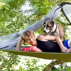 Un chico sonríe mientras su perro se asoma por encima de él en una hamaca con mosquitera, disfrutando del aire libre.