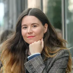 Johanna Hedman, med sitt långa, vågiga hår och en rutig kavaj, ler mot betraktaren.