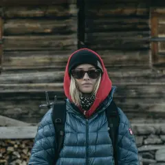 Kobieta w czerwonej bluzie z kapturem i czarnej czapce, w okularach przeciwsłonecznych, z plecakiem, przed drewnianą chatą.