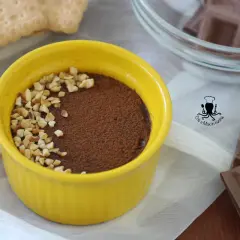 Budino al cioccolato in ciotola gialla, cosparso di cacao e nocciole tritate. Accanto, biscotti e cioccolato.