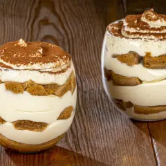 Due bicchieri di tiramisù senza uova, stratificati con crema e savoiardi, spolverati di cacao, su un tavolo di legno.